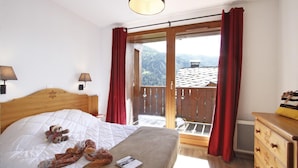 Un dormitorio con una cama, un aparador de madera, una mesita de noche, una lámpara y vistas a un balcón con cortinas rojas.