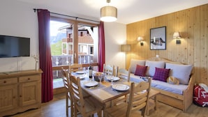 Un comedor con una mesa de madera dispuesta para cuatro personas, un sofá con cojines, un televisor y vistas a un paisaje montañoso a través de una puerta corredera.