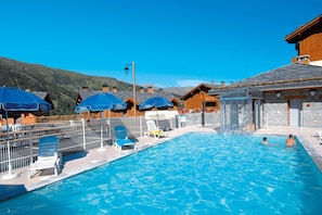 Una piscina con lettini e ombrelloni, circondata da edifici in legno e con montagne sullo sfondo.
