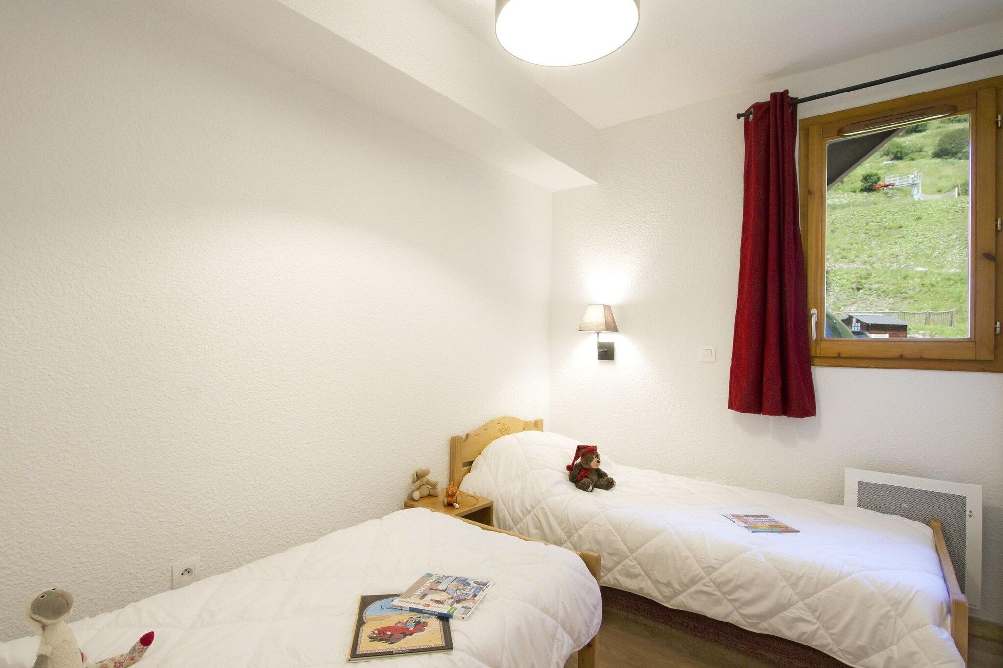Une chambre avec deux lits, une fenêtre donnant sur un paysage verdoyant, une lampe fixée au mur et un rideau rouge.