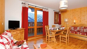 Um quarto aconchegante com piso de madeira, uma televisão sobre um móvel de madeira, uma mesa de jantar com cadeiras e um sofá com capa estampada.