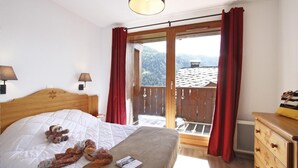 Quarto com cama, cómoda de madeira, mesa de cabeceira, duas luminárias de parede e janela com cortinas vermelhas.