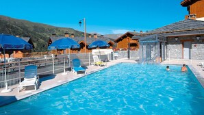 Piscina com espreguiçadeiras e guarda-sóis, cabanas de madeira e montanhas ao fundo.