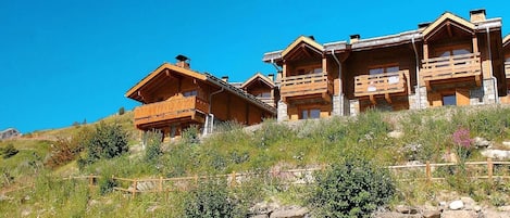 Una hilera de cabañas de madera con balcones, ubicadas en una colina bajo un cielo azul despejado.