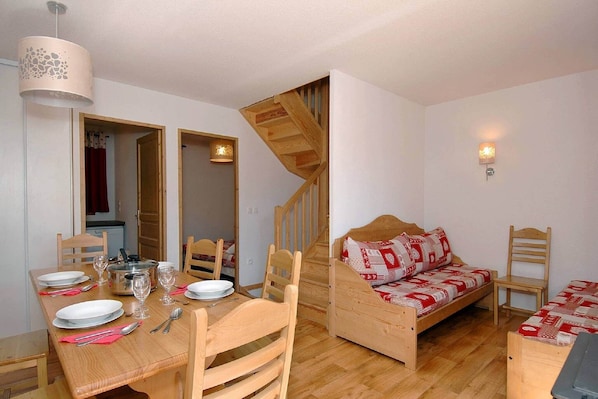 A compact room with a dining table set for four, a wooden bed with a patterned blanket, a wooden staircase, and a wall-mounted light fixture.