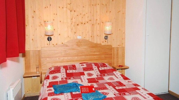Quarto com cabeceira de madeira, duas luminárias de cabeceira, colcha com padrão vermelho e branco e cortina vermelha.