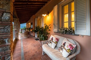 A covered outdoor seating area with wicker furniture, a stone wall, and a row of windows with shutters.