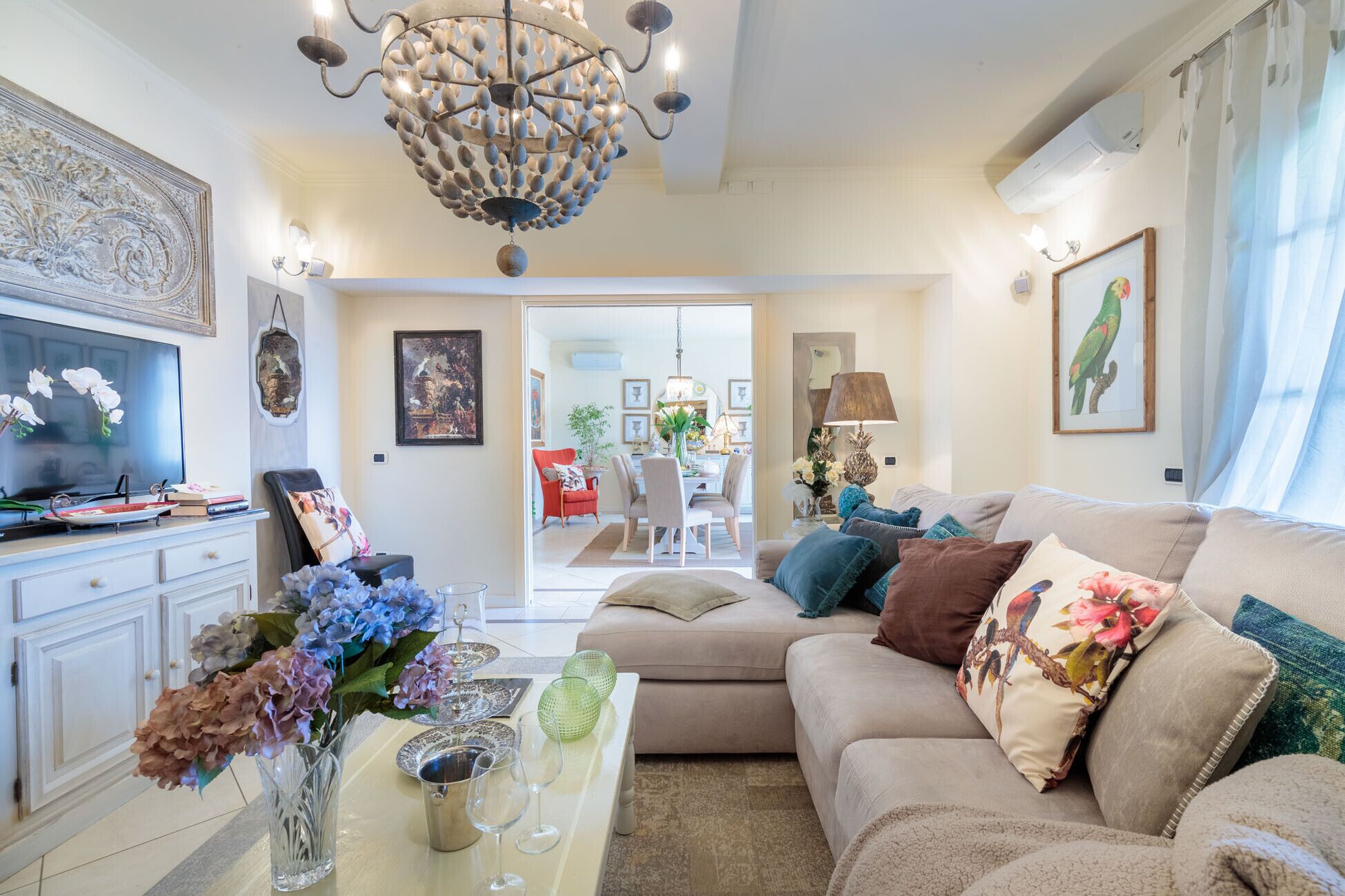 Un salon avec un canapé beige, un téléviseur et un lustre. On aperçoit une salle à manger à travers une porte ouverte.