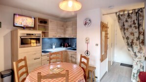 Una cocina compacta con armarios de madera, horno integrado, mesa de comedor con mantel a cuadros y un televisor en la pared.