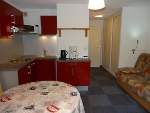 A compact kitchen with red cabinets, a sink, and a coffee maker. There's a bed with a patterned bedspread and a sofa with a patterned cover.