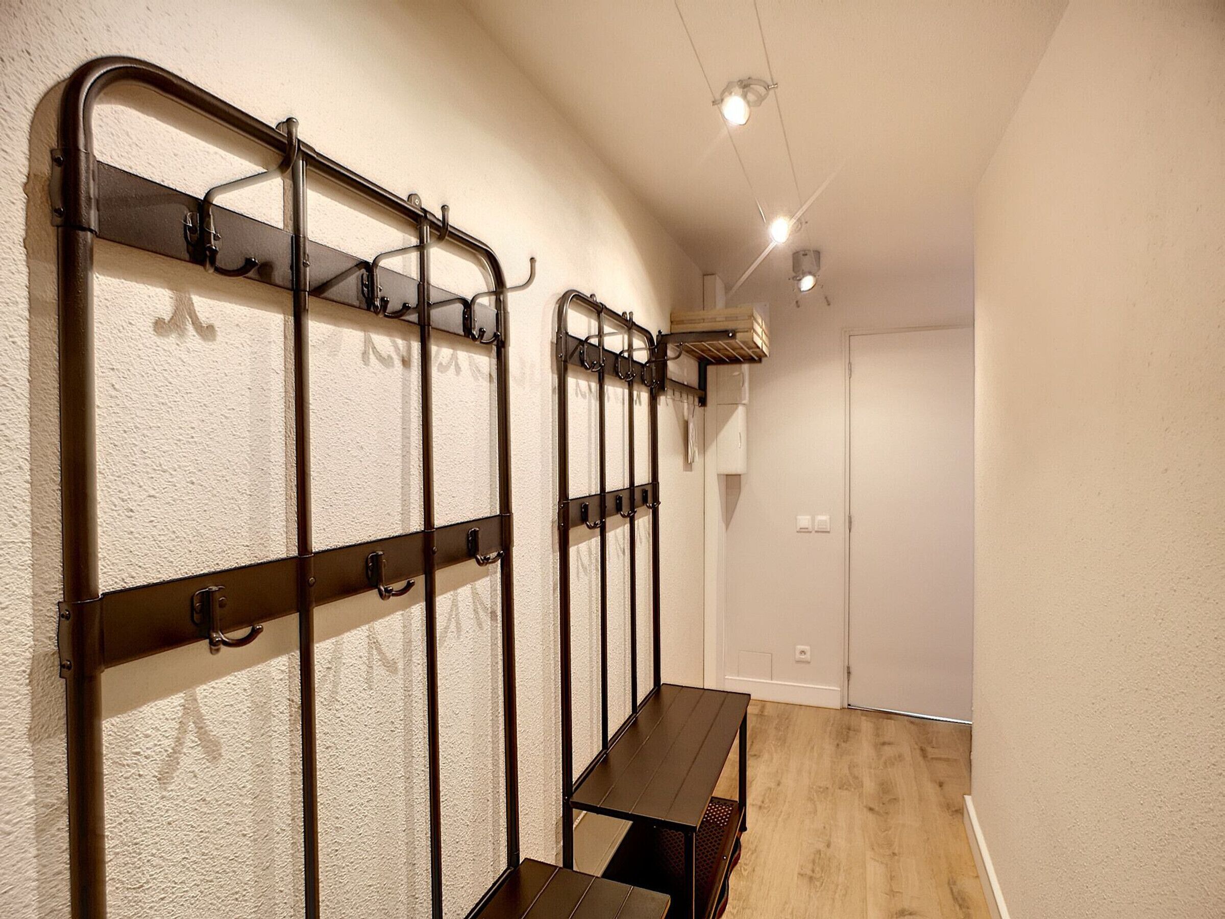 Un couloir bordé d’une rangée de bancs en métal et de crochets fixés au mur, un banc en bois au centre, et une porte blanche au fond.