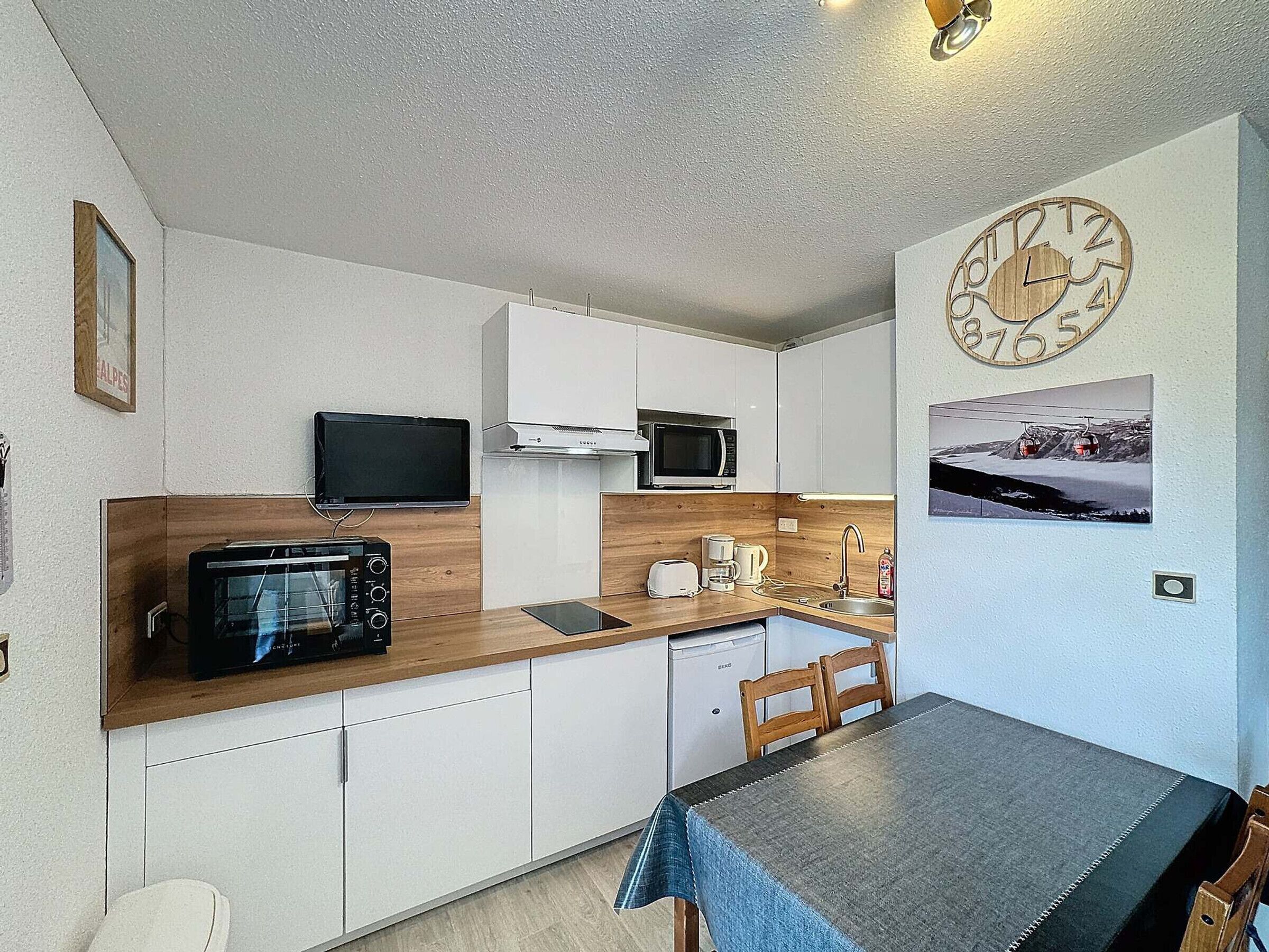 A compact kitchen with white cabinets, a microwave, a toaster, and a sink. There's a wooden countertop, a dining table with chairs, and a wall clock.