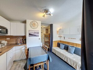 A compact kitchen with a microwave, sink, and wooden countertop. A dining table with chairs is positioned in front of the kitchen. There is a bed with a headboard and pillows in the adjacent room.