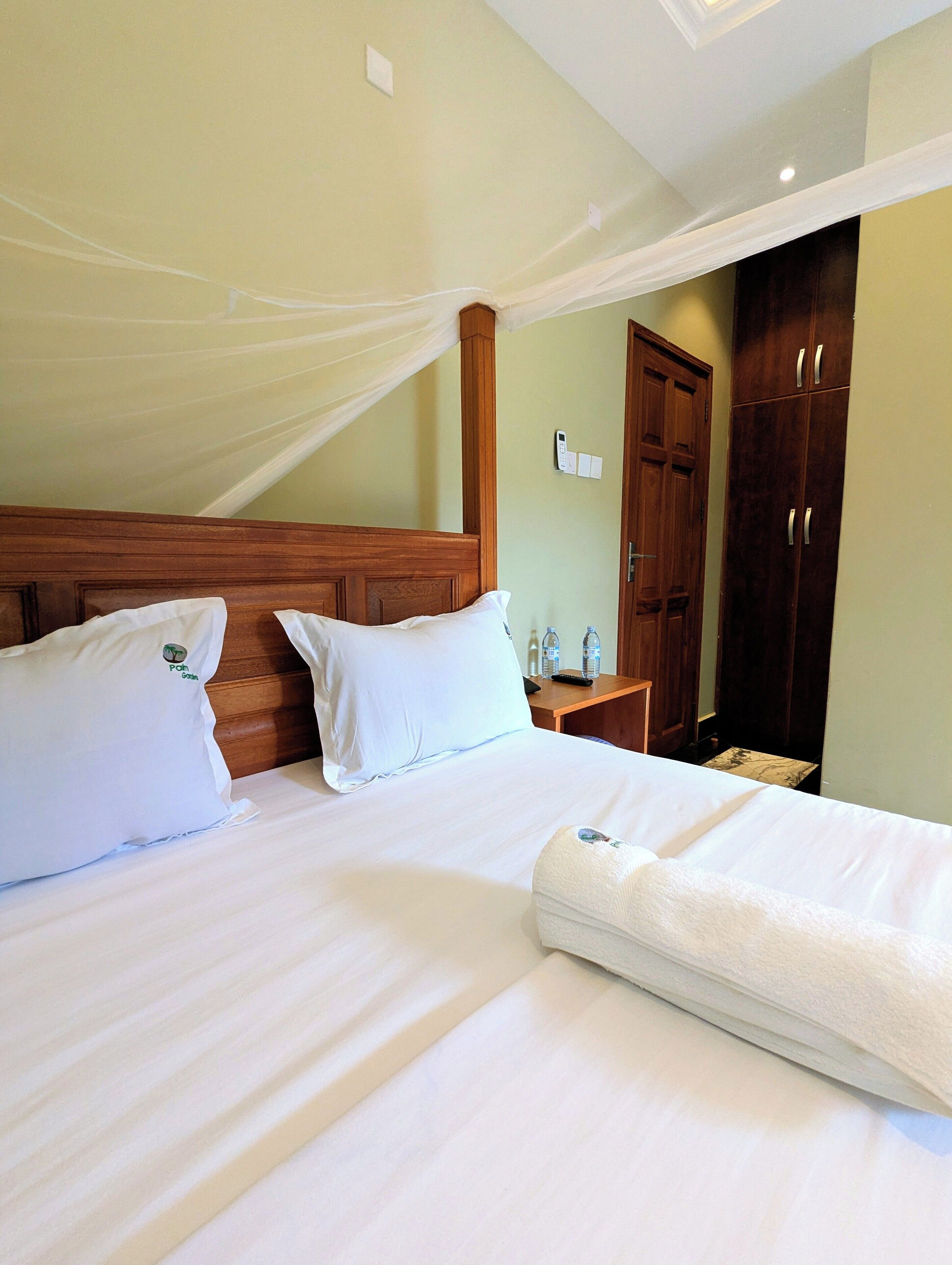 A hotel room with a large bed, wooden headboard, and a canopy. There is a nightstand with a remote control and a bottle of water. A wardrobe is visible in the background.
