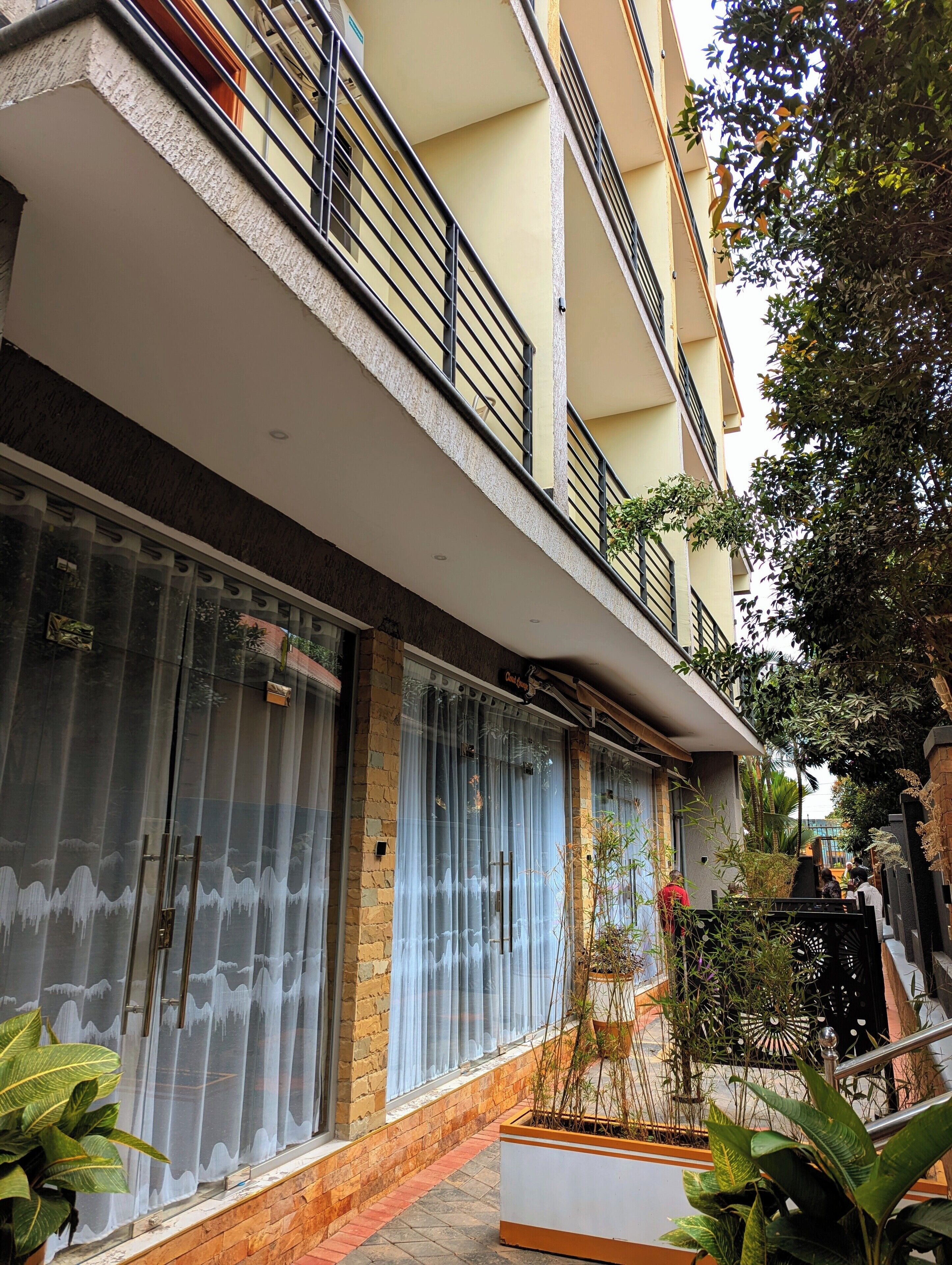 Ein mehrstöckiges Wohngebäude mit Balkon, großen Fenstern mit Vorhängen und einer kleinen Gartenanlage mit Topfpflanzen.