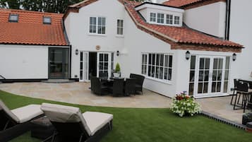 A two-story house with a patio area featuring a dining table and chairs, a small garden, and a lawn.
