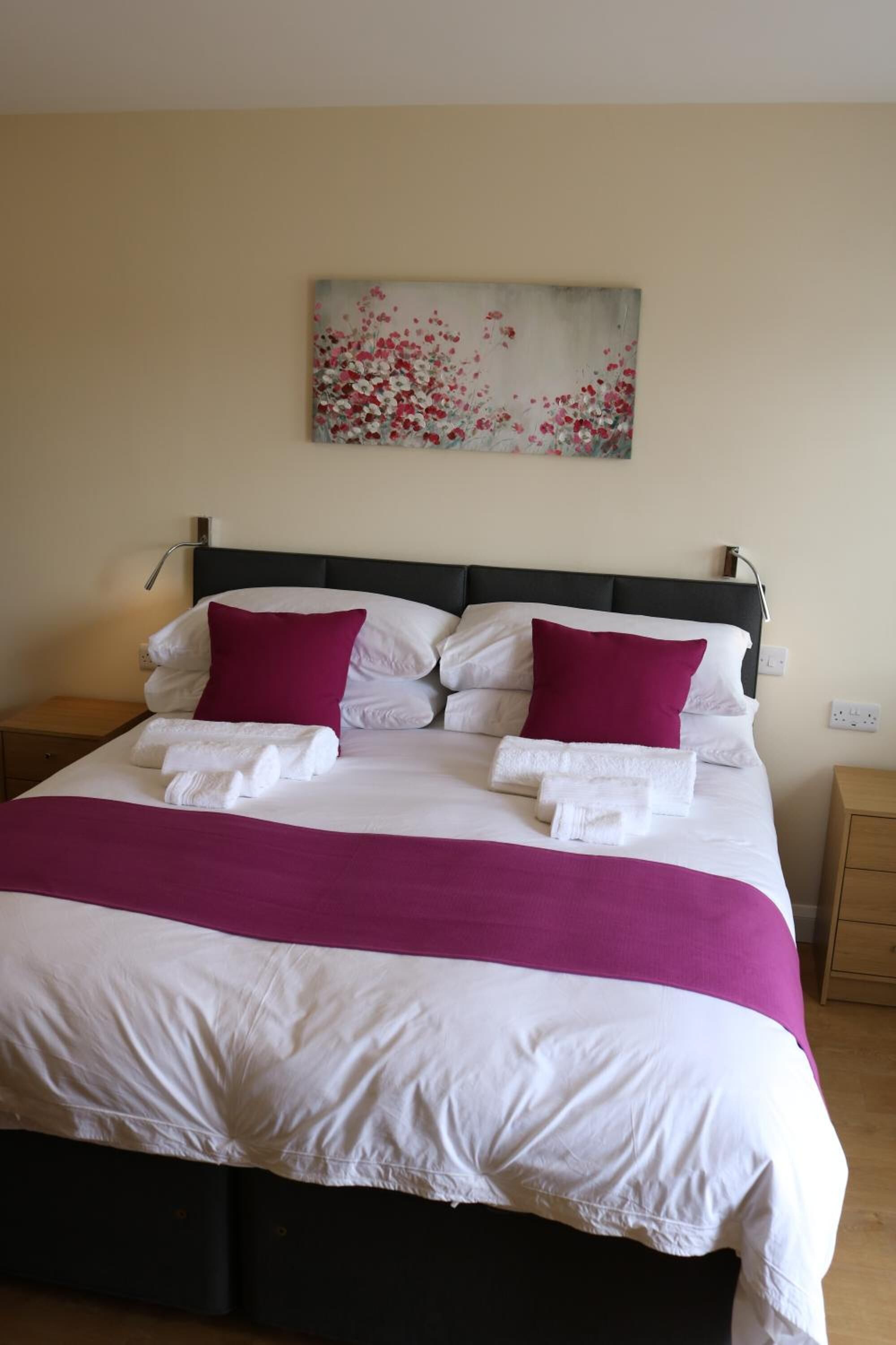 Una cama bien tendida con ropa de cama blanca y morada, un cabecero negro y un cuadro con flores en la pared.