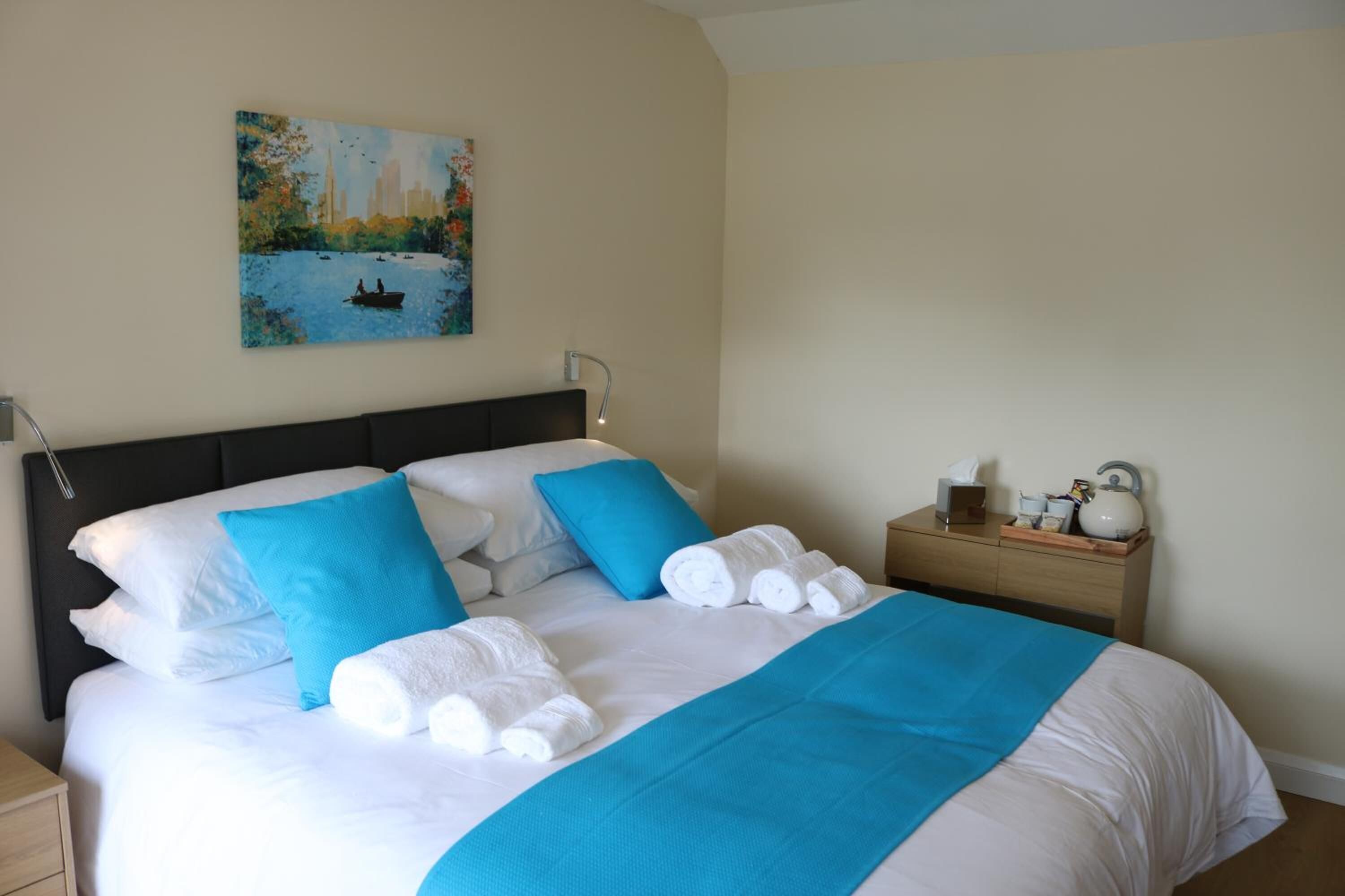 A neatly made bed with white linens and blue accents, a bedside table with a lamp and a small decorative item, and a painting of a cityscape on the wall.