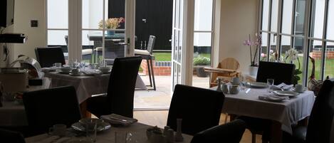 A dining area with tables set for four, a television mounted on the wall, and a view of the outdoor patio through large windows.
