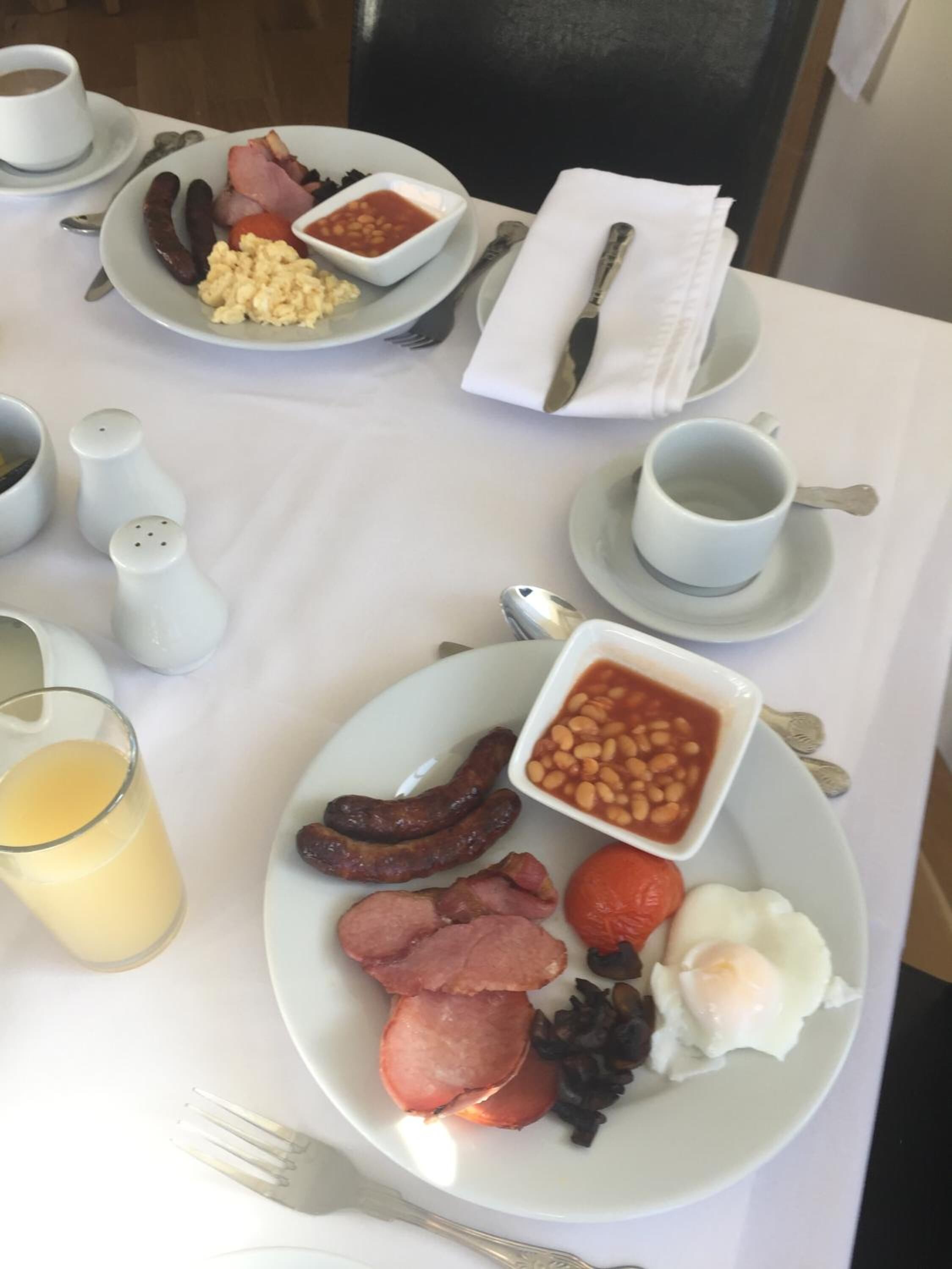 A breakfast setting with plates of sausages, beans, bacon, eggs, and toast, along with cups of coffee and a glass of orange juice.