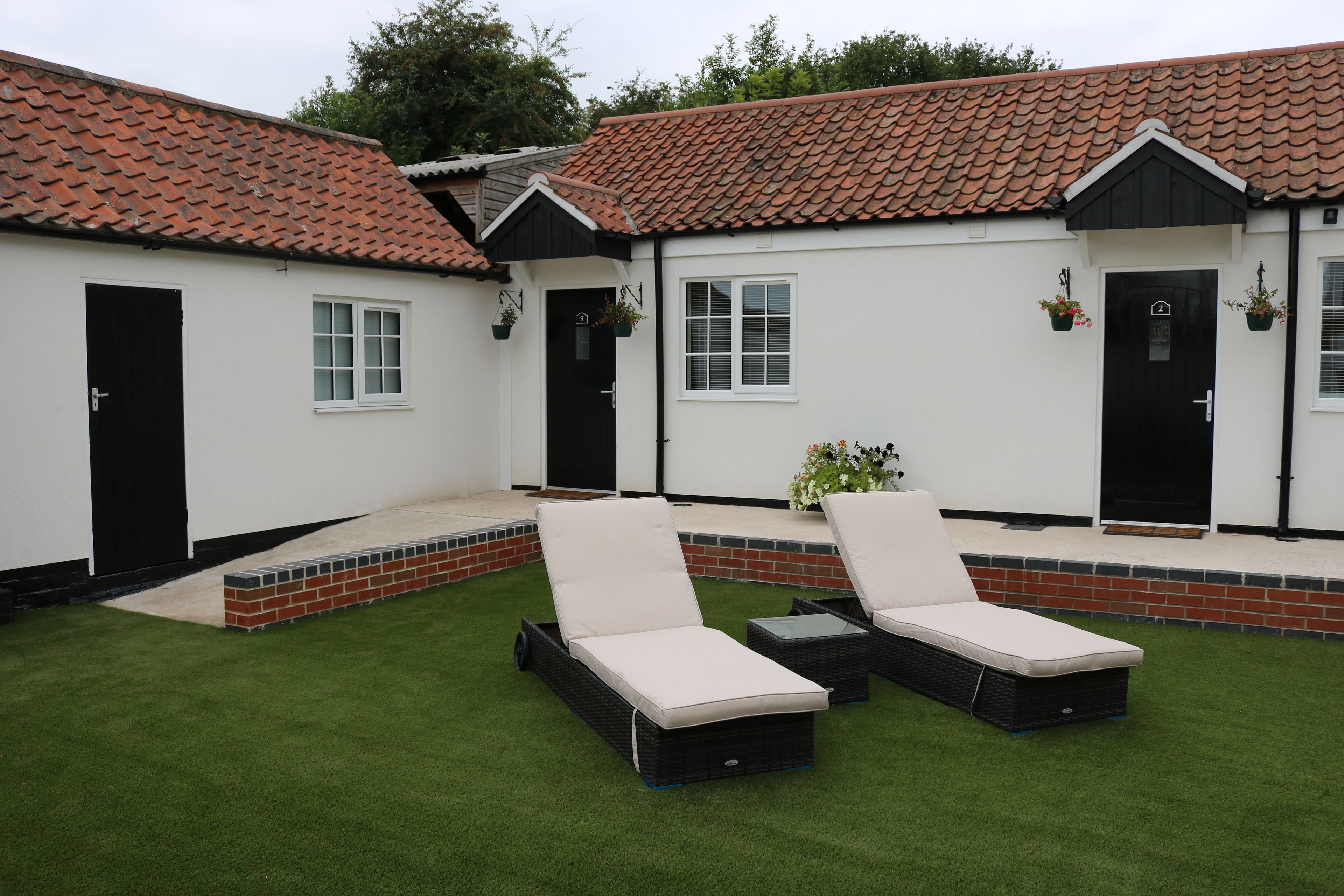 A white building with a red-tiled roof, two black doors, and a small patio area with two black lounge chairs and a table.