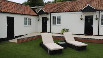 A white building with a red-tiled roof, two black doors, and a small patio area with two black lounge chairs and a table.