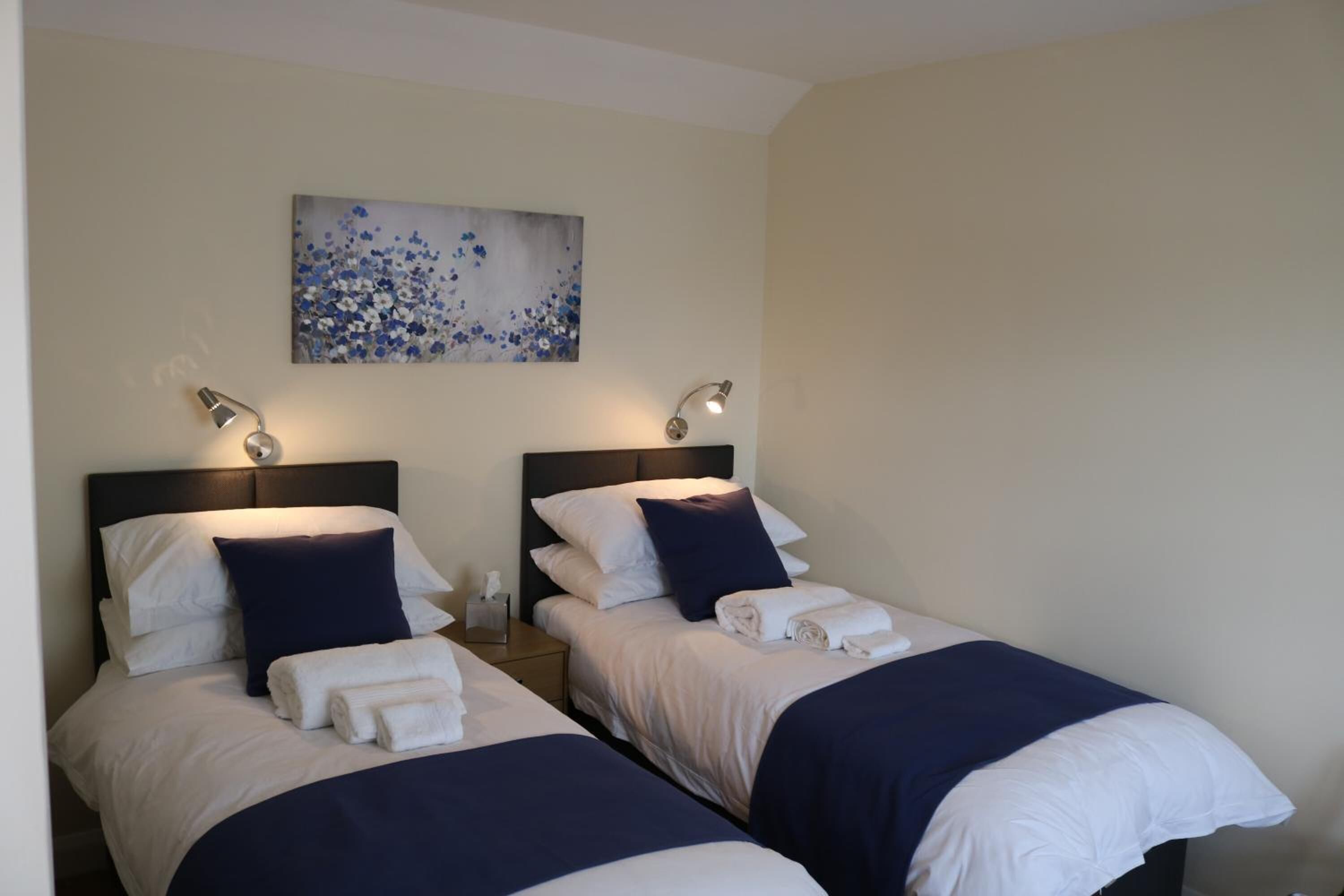 A hotel room with two beds, each with white linens and dark blue bedding. There is a painting on the wall above the beds, and two bedside lamps.