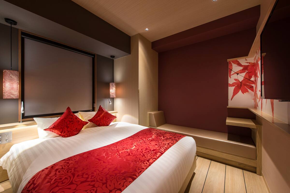 A modern hotel room with a large bed, red and white bedding, a wooden bench, and a red maple leaf artwork on the wall.