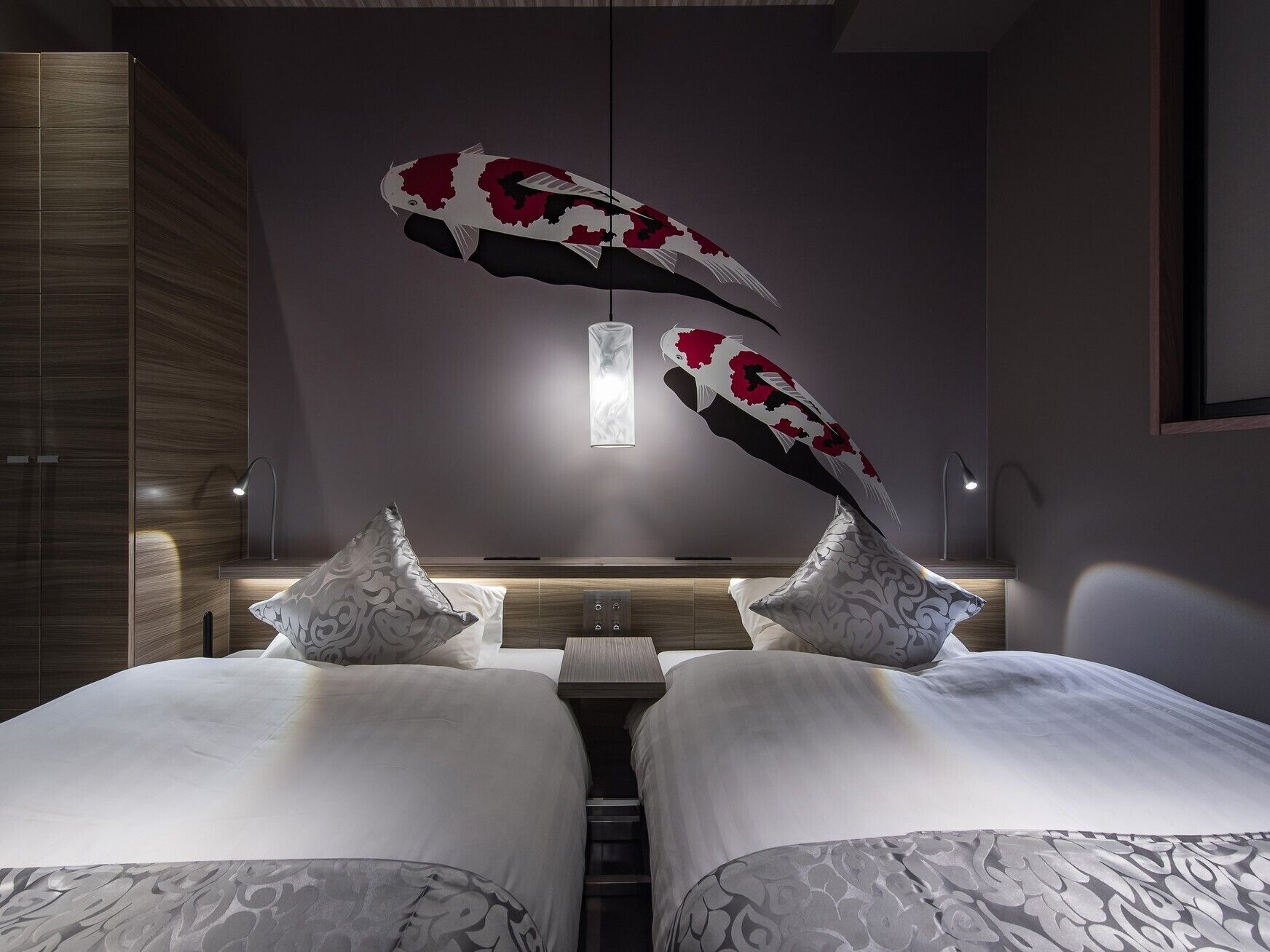 A modern hotel room with a central bed, two decorative pillows, a hanging light fixture, and a wooden wardrobe.