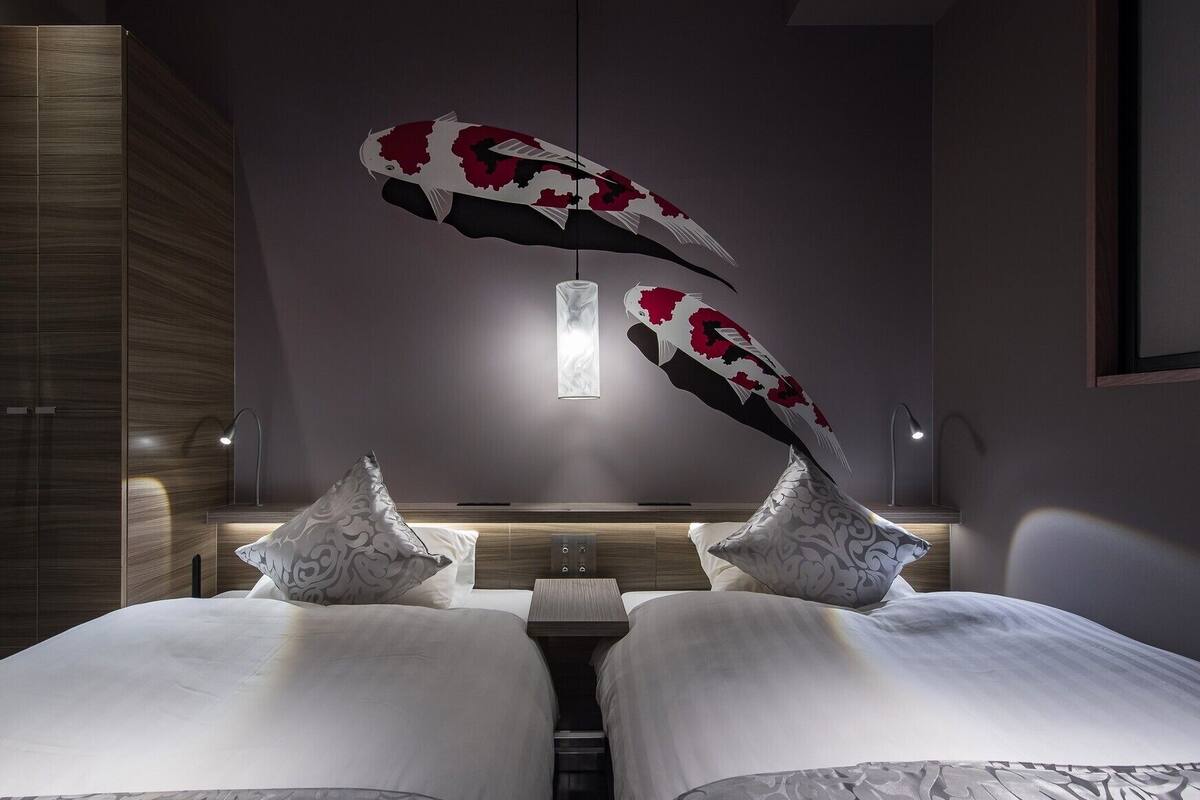 A modern hotel room with a central bed, two decorative pillows, a hanging light fixture, and a wooden wardrobe.
