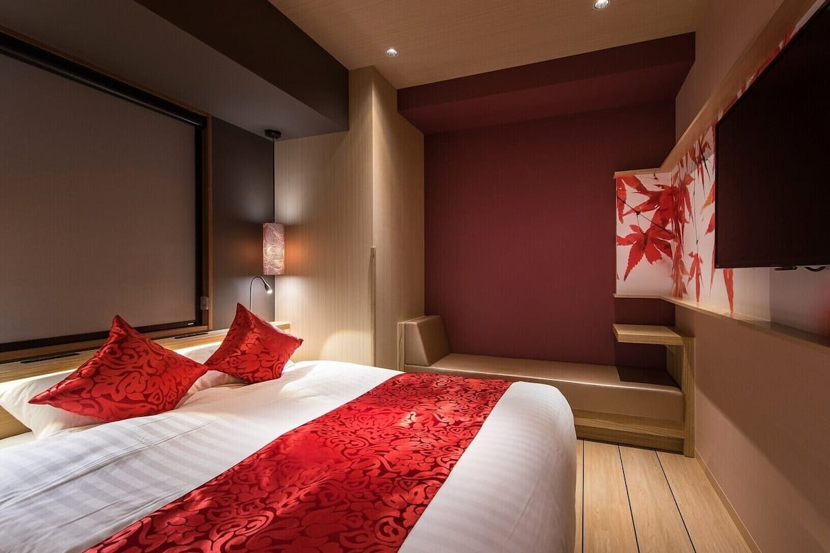 A modern hotel room with a large bed, red patterned bedspread, and a seating area with a red leaf wall art.