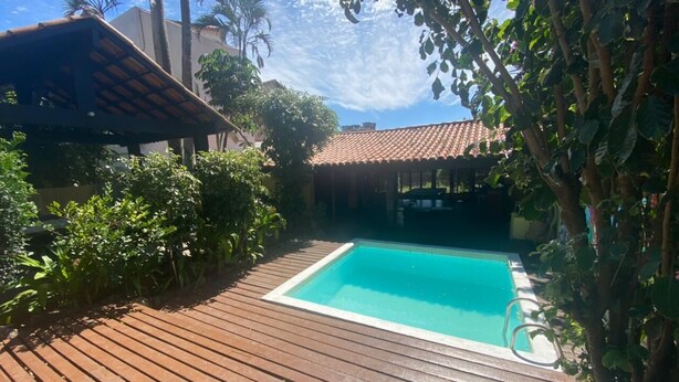 A wooden deck with a swimming pool, surrounded by greenery and a tiled-roof structure. - Beautiful and Comfortable House for up to 15 People in Arraial do Cabo (Location: Vacation Rental) (Arraial do Cabo)