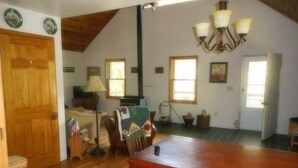 A spacious room with a wooden dining table, a sofa, a chair, and a lamp. There are two windows providing natural light and a door leading to another room. - Mountain views, total seclusion Southern Bedford County, PA - Big Discounts! (Clearville)