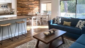 A modern kitchen with a wooden accent wall, a dining area with white chairs, and a blue sofa with patterned cushions.