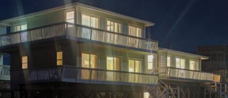 A multi-story beachfront house with a balcony on each floor, featuring a wooden deck and a staircase leading to the upper levels.