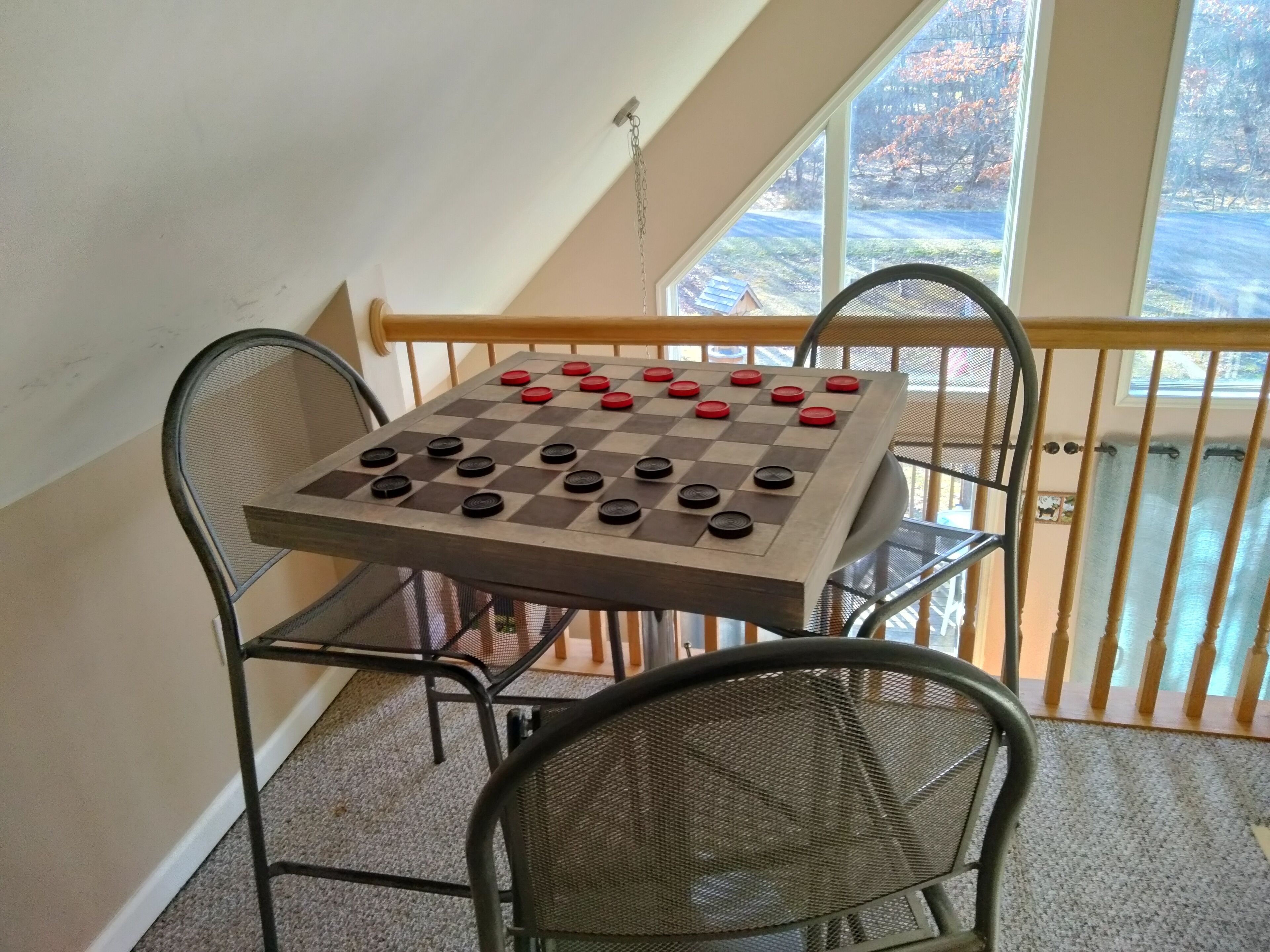 A chessboard on a table with chairs, set on a balcony with a view of trees and a house.