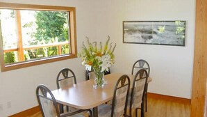 Un coin repas avec une table ronde, six chaises, un vase de fleurs, une fenêtre et un tableau encadré au mur.