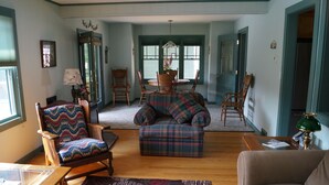 A cozy living room with a wooden floor, a plaid sofa, a wooden armchair, a glass coffee table, and a rug.