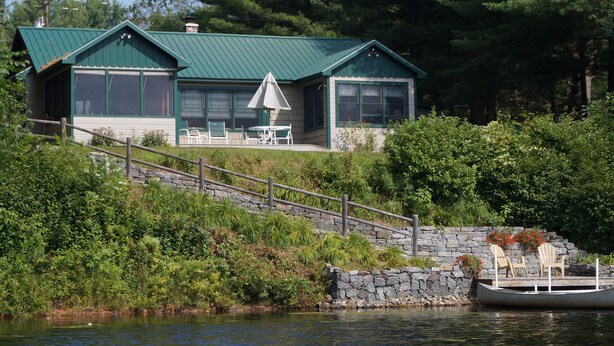 A lakeside cabin with a stone staircase leading to a deck, a boat docked at the water's edge, and outdoor seating.