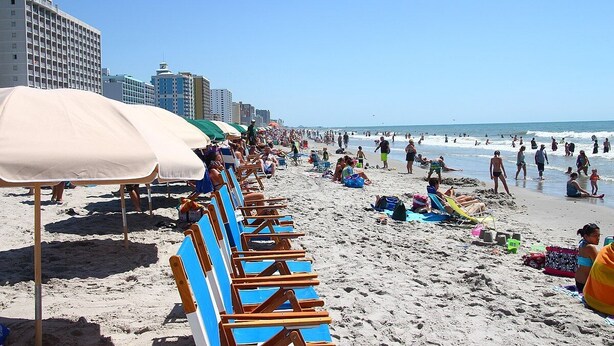Una playa con hileras de sillas azules y sombrillas, una orilla arenosa y edificios al fondo.