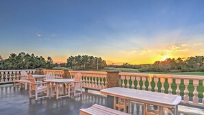 Un patio al aire libre con mesas y sillas de madera, un balcón de piedra y vistas al atardecer sobre un campo de golf.