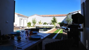 Een terras buiten met een houten eettafel, een barbecue en een wit gebouw met pannendak op de achtergrond.
