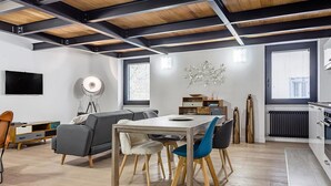 A modern living room with a dining area, wooden beams on the ceiling, a flat-screen TV, and a radiator.