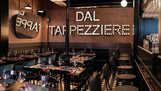 A restaurant interior with brick walls, illuminated signage, and a checkered floor. Tables are set with wine glasses and plates.