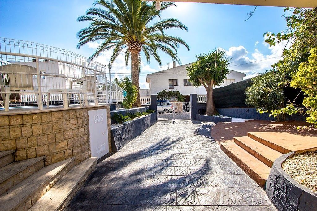 A stone pathway leading to a gated entrance with a palm tree and a white building in the background.