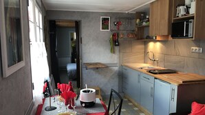 Une cuisine compacte avec un plan de travail en bois, un évier et un four à micro-ondes. On y trouve un coin repas avec une table dressée pour deux personnes, une nappe rouge et une chaise. La cuisine est dotée de placards bleu clair et d’un sol carrelé.
