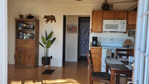 A cozy kitchen with wooden cabinets, a microwave, and a coffee maker. There's a dining area with a table and chairs, and a bookshelf filled with books and decorative items.