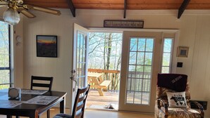 A cozy room with a wooden ceiling, a dining table with chairs, a cushioned chair with a pillow, and a large window with a view of the outdoors.
