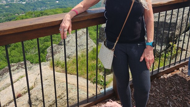A person standing on a wooden deck with a metal railing, overlooking a mountainous landscape.