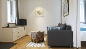 Una sala de estar moderna con un sofá gris, una pequeña mesa de madera con mantel de encaje, un televisor en un mueble de madera y un cuadro enmarcado en la pared.